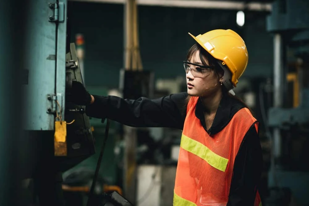 Female industrial engineer wearing a white helmet while standing in a heavy industrial factory behind she looking of working at industrial machinery and check security system setup in factory.