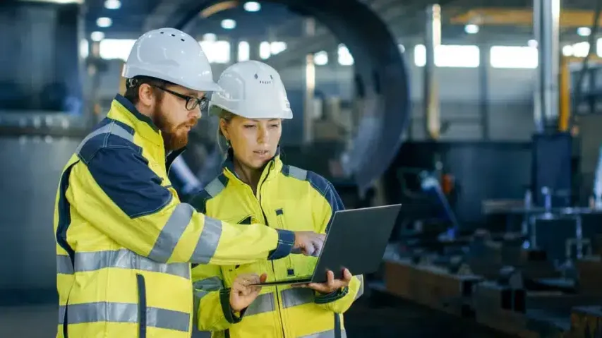 Team members in high-visibility jackets and helmets discussing Behavior-Based Safety strategies on-site using a laptop.