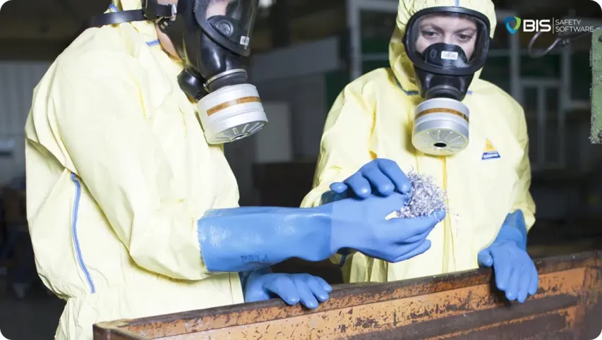 Two workers in yellow hazmat suits, blue chemical-resistant gloves, and full-face respirators examining a handful of metal shavings in an industrial setting.