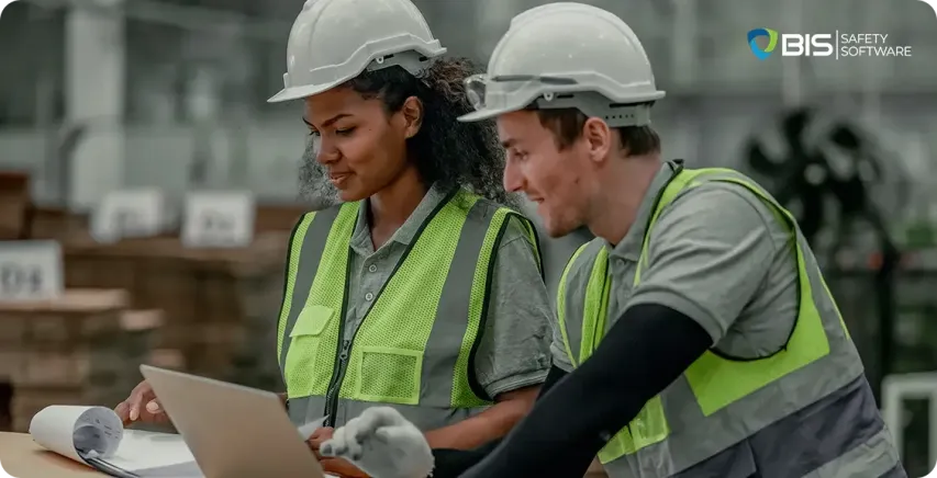Two workers in safety vests and hard hats collaborate on a laptop in a warehouse setting, highlighting the role of communication, digital tools, and mental well-being in maintaining a safe and productive workplace.