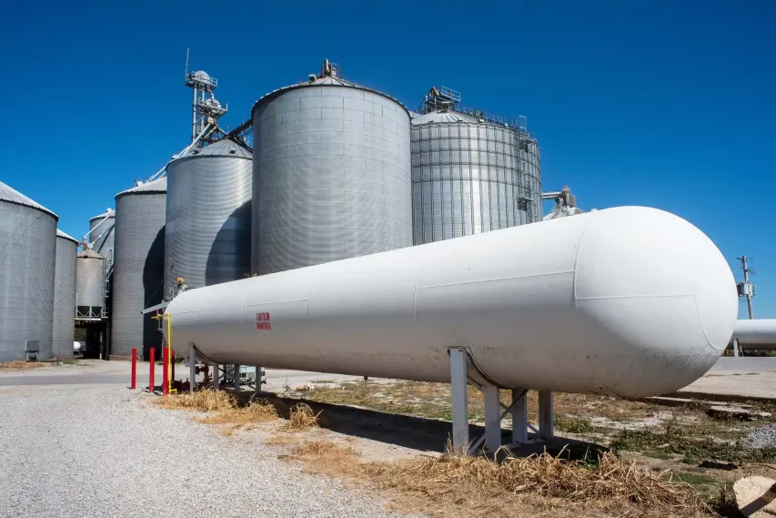 Bulk ammonia storage tank stands at an agricultural facility, with silos and safety bollards in view.