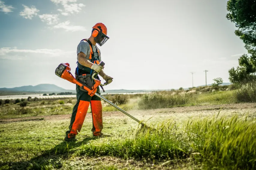 Landscaping worker using a string trimmer while wearing full PPE including visor, gloves, and high-visibility gear.
