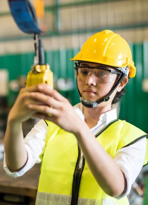 Waist up female worker with safety helmet press control panel for lifting up or down of power trolley crane in factory warehouse. Asian woman controls crane beam in manufacturing facility.