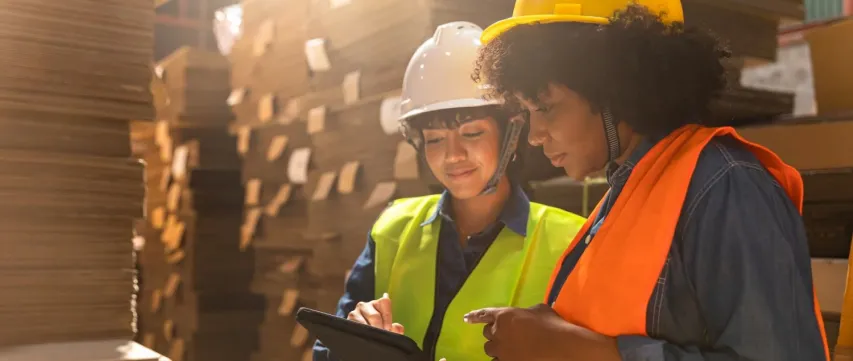 Engineer asian and african woman wearing safety helmet and vest holding clipboard and take note on the paper in the automotive part warehouse.Products and corrugated cardboard. Factory for the manufacture and processing of paper.